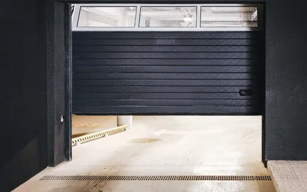 Partially open black industrial roller shutter door inside a loading bay with concrete floor and drainage channel